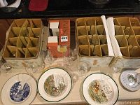 Overview of two padded storage cases with multiple clear glass stemware pieces with gold rims, and three decorative plates placed in front.