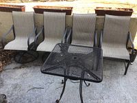 Photo showing four mesh patio chairs arranged side by side against a wall with black metal frames and beige mesh seats, with black metal square mesh table in front.