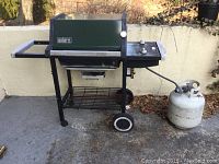 Full side view of the Weber 8127 propane grill on black metal frame with wheels and propane tank attached, shown outside with concrete floor and stone wall background.