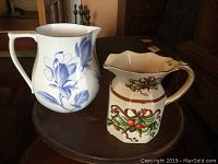Two Tiffany porcelain pitchers on a round table, one with blue floral design, the other with holiday ribbon and greenery motif