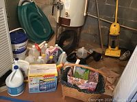 View showing assorted bottles of cleaning liquids, two baskets (wooden with liner, plastic), bungee cords, and plungers arranged on floor near water heater and yellow vacuum cleaner.