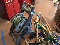 Photo showing plastic and metal mobile hose reel with a green garden hose coiled, surrounded by various wooden and metal handled garden tools and cleaning tools on garage floor.
