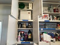 Open white cabinets displaying a white bread maker, several spray bottles and cleaning liquids such as window cleaner, dish soap, and disinfectants.