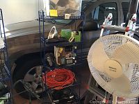 Full view of the shelving unit with multiple shelves holding coiled cords, boxes, fan, heater, and other items against garage background.