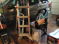 Photo of Little Giant Helium step ladder standing upright in garage, visible ladder base wheels and folded stools in background.