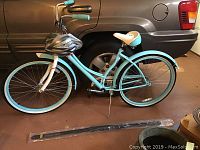 Side view of light blue Schwinn ladies cruiser bicycle with helmet on handlebars and Bodyblade exercise device on floor near front wheel.