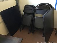 Two stacked black Cosco folding card tables leaning against the wall with a stack of folded metal chairs next to them.