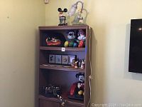 Full view of bookcase with various Disney Mickey Mouse collectibles displayed including plush figures, ceramic figurines, clock and phone.