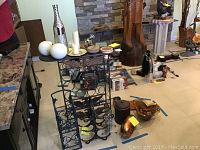 Wide angled photo showing the large metal wine rack, smaller wine rack, copper cups, art glass, candles, and coasters arranged on floor with some items on table near fireplace.