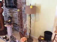 Photo showing faux wood floor lamp with beige drum shade, and faux wood vase containing tall twisted faux wood sticks and white branches, with peach artificial flower near base.