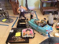 Wide shot showing multiple Elvis collectible items arranged on the floor, including figurines, pot holders, toy car, plaques, and spoon.