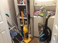Closet showing vacuums including yellow upright vacuum, cleaning tools, step ladder, and laundry area.
