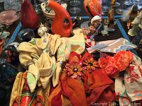 Photo shows assorted cloth placemats and napkins in beige, red, and patterned designs spread on a surface with various glass figurines and paperweights, including a glass cockatiel and colorful glass fish. Also includes oil and vinegar containers and decorative jar.