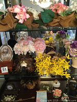 Shelf with six sets of assorted faux flowers including pink hydrangeas, yellow flowers, and others, along with porcelain flower statues, decorative mask, and clocks.