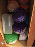 Photo of kitchen cabinet containing multiple plastic food storage containers with colored lids (purple, green, orange) stacked inside a wooden cabinet.