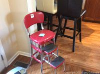 Front side angle view of red metal kitchen chair placed on wooden floor near dining table. Chair has curved backrest, red glossy seat, metal tubular legs and built-in two-step ladder.