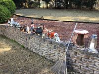 Wide view showing multiple small outdoor yard statues including rabbit, rooster, watering can, frog, and metal bull on a garden retaining wall with broom and lantern at one end