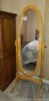 Full front view of an oval standing mirror with a light wood frame, turned supports, and a carpeted floor with bedroom furniture in background.