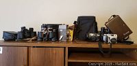 Wide shot on wooden cabinet showing cameras, binoculars and cases lined up horizontally