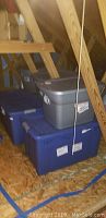 Four plastic storage bins (two blue, one gray) stacked under attic rafters
