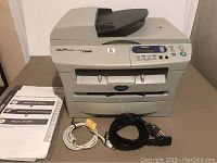 Front and top view of Brother DCP 7020 laser printer with control panel, paper output tray and document feeder visible. Accessories and manuals placed beside the printer.