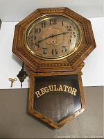 Full view of the clock face with wooden octagonal frame and drop bottom door labeled 'REGULATOR', showing pendulum and key placed beside it.