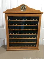 Front view of the wooden golf ball display case showing seven shelves with wooden pegs (tees) for holding golf balls, glass door, and decorative metal plaque at the top.
