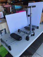 Side view of Leg Magic exercise machine showing dual black foot platforms on rails, a central handlebar with foam grips labeled 'LEG MAGIC'.