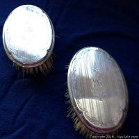 Two oval, sterling silver backed brushes with white bristles on dark fabric. Visible intricate engraving and monogram on silver backs.