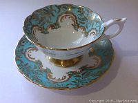 Angle view of Royal Stafford cup sitting on saucer showing scalloped edges, gold footed base, turquoise and gold filigree decoration.