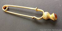 Full view of the gold colored Scottish kilt pin showing the long pin body and thistle top with amber stone.