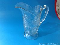 Clear molded glass pitcher with embossed countryside and hunting dog scenes, shown standing upright against blue background.
