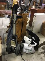 Full golf bag with golf clubs, shoes in a white bag, gray nylon jacket draped over golf cart, and golf cart stand.
