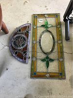 Two stained glass panels lying on the floor, one rectangular with green leaf motif and one semi-circular panel with rose design.