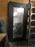 Full view of large wood framed floor mirror leaning against wall, showing black wide frame and full-length glass.