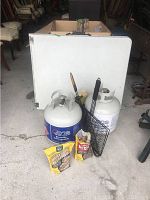 Photo showing two propane tanks, folding table folded up behind, bags of wood chips, and grilling baskets.