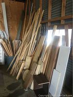 View showing large assortment of long wooden planks leaning against a wall, with several flat and round wood pieces on the floor next to them.