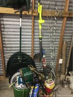 Photo showing Ryobi Easy Edge weed trimmer standing upright next to shovel and green plant watering can. Also visible are plant stands with spiral design, garden pegs, and garden tools in plastic bucket.