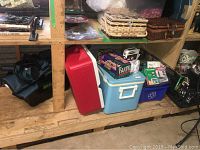 Shelf with two Coleman coolers (red and blue), woven wicker baskets, portable alert signal, board game Bandu, small camera, and storage bins.
