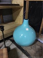Photo of large blue vase with bulbous shape and narrow neck on plywood surface against wooden wall.