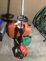 Photo of the gas weed trimmer standing upright with an orange gas canister and green gas container behind it on a concrete floor.