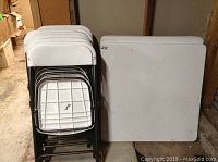 Two folded square white plastic card tables with black metal legs next to a stack of seven folded white plastic chairs with black metal frames, stored indoors on a concrete floor.