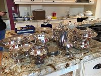 Front view of various silver-plated serving pieces including covered bowls, a tea pot, pedestal trays, and sugar bowl arranged on a granite countertop.