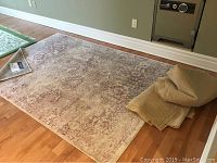 Full view of the rose and beige colored area rug on hardwood floor with some folded rugs piled at one end.