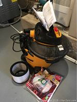 Shop-Vac wet/dry vacuum tank with cords and owner's manual on top, cylindrical filter and vacuum bags on the floor beside it.