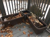 Wide view showing two rectangular brown plastic flower bed planters and square metal wicker planter on an outdoor deck with dry leaves.
