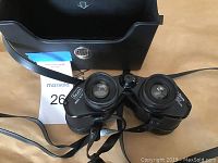 Front view of black Sears 7x35 binoculars with attached neck strap placed in front of black carrying case with metal clasp.