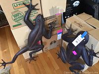 Two metal frog sculptures shown against cardboard boxes on a wooden floor. Large frog on the left, smaller frog on the right.