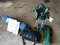 Photo showing the folded green camping chair, blue sunshade bag with owner's manual, and black fanny pack placed on a concrete floor.