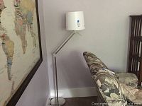 Floor lamp with metal adjustable telescoping arm, white cylindrical shade, placed against a wall near an armchair and a large wall map.